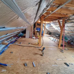 Montaż izolacji termicznej poddasza z widocznymi rurami instalacji wodnej i elektrycznej oraz wkrętarką na podłodze z płyty OSB.