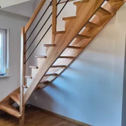 Drewniane schody z czarnymi metalowymi poręczami w jasnym wnętrzu, widoczne parkiet i okno.