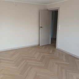 Horyzont Sebastian Zapora - Jasne parkiety ułożone w jodełkę w pustym pokoju z otwartymi, szarymi drzwiami. Widoczne listwy przypodłogowe i wystające kable z sufitu.