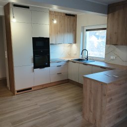 Horyzont Sebastian Zapora - Nowoczesna kuchnia w odcieniach bieli i drewna z czarnymi akcentami, widok na okno i zabudowę kuchenną z piekarnikiem i płytą. Podłoga drewnopodobna.