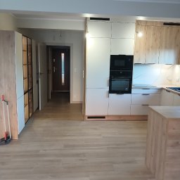 Horyzont Sebastian Zapora - Nowoczesna kuchnia z zabudową w odcieniach bieli i drewna, z widocznym sprzętem AGD i wyspą kuchenną. Jasne panele podłogowe i otwarte przejście do przedpokoju.