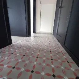 Wzorzysta podłoga z płytek w odcieniach szarości, czerwieni i czerni, widziana z perspektywy niskiego kąta, prowadząca przez otwarte, ciemne drzwi do jasnego pomieszczenia.