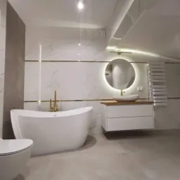 Nowoczesna łazienka z wolnostojącą wanną, umywalką nablatową podświetlaną okrągłym lustrem i grzejnikiem drabinkowym, wykończona białymi płytkami imitującymi marmur z akcentami złotych pasków.