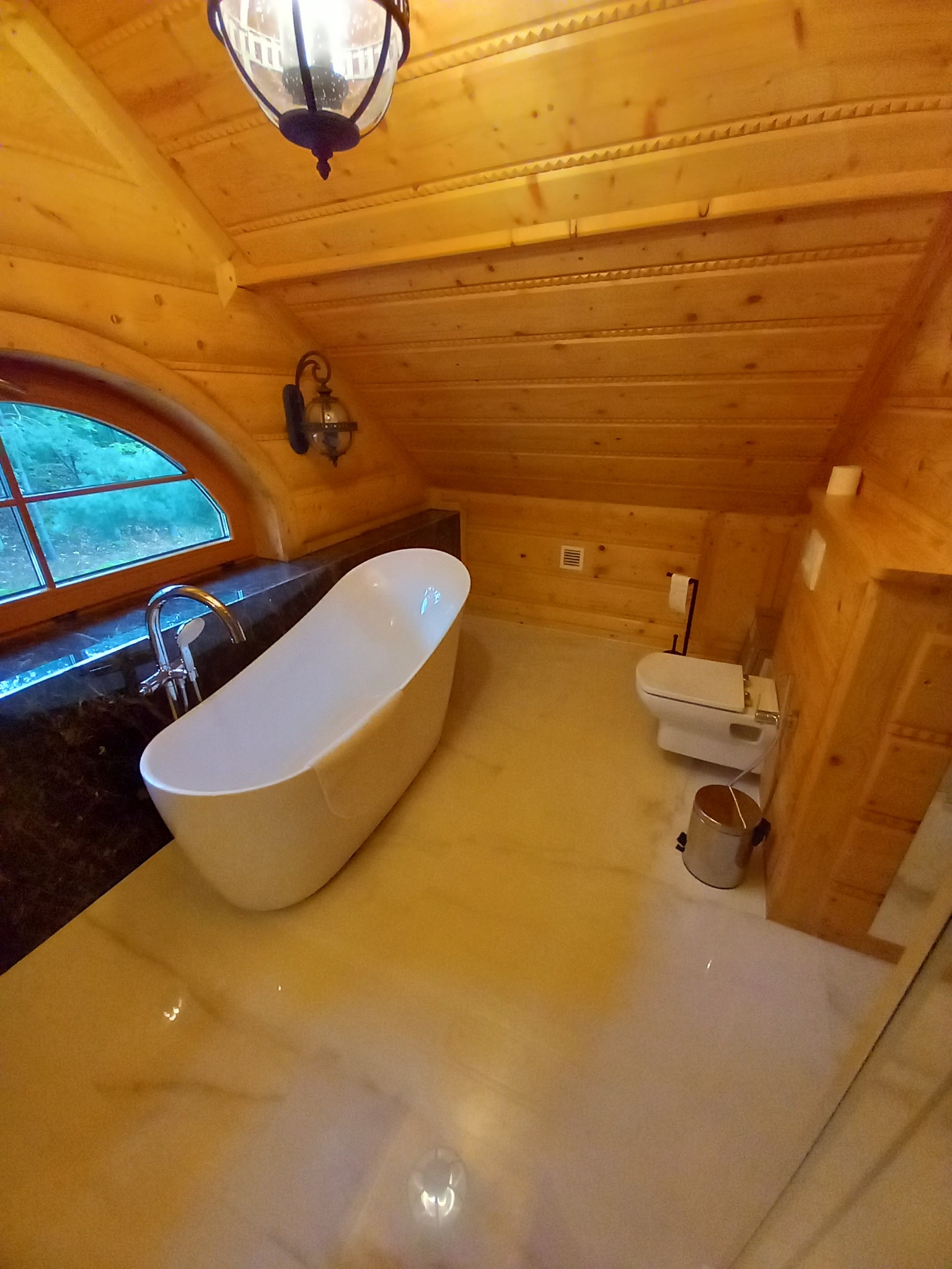 Drewniana łazienka na poddaszu z wolnostojącą wanną, oknem w kształcie łuku i marmurowym blatem, oświetlona lampą sufitową i kinkietem.