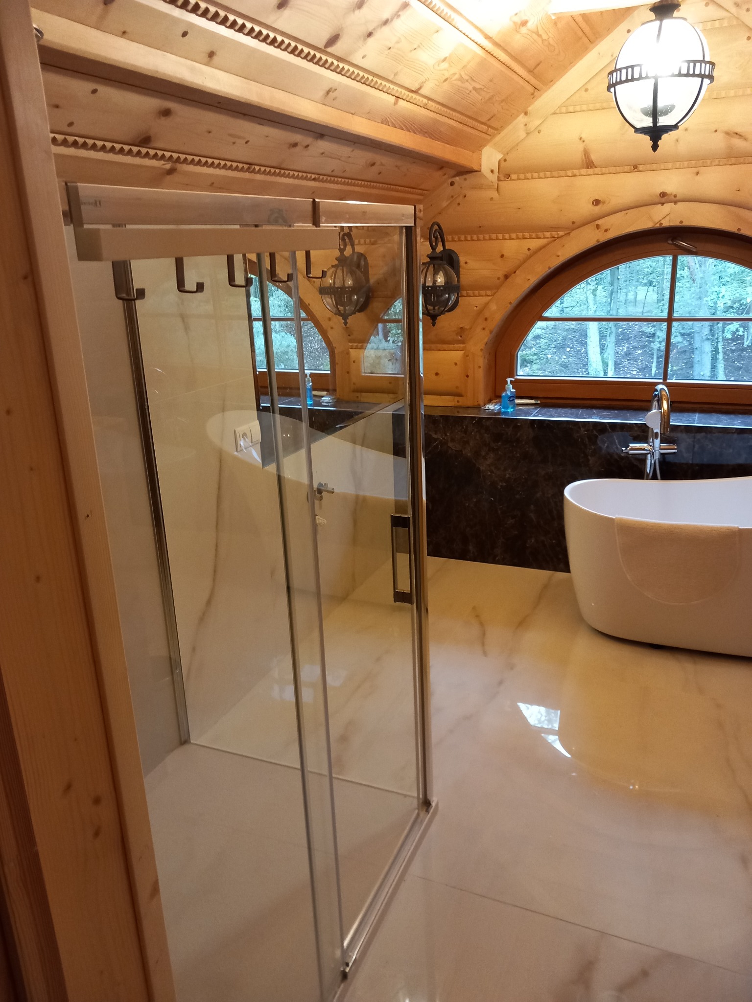 Wykończona łazienka w stylu góralskim z drewnianym sufitem, przeszkloną kabiną prysznicową z uchwytami na ręczniki i wolnostojącą wanną przy oknie.