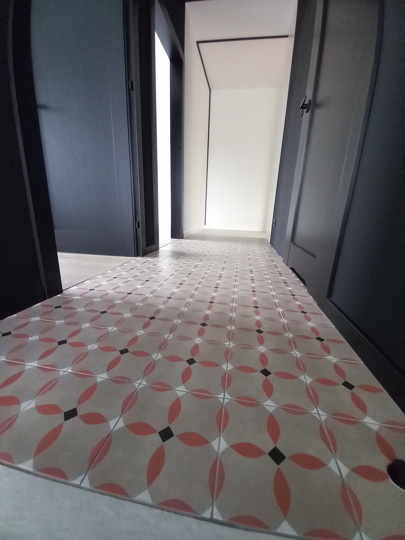 Wzorzysta podłoga z płytek w odcieniach szarości, czerwieni i czerni, widziana z perspektywy niskiego kąta, prowadząca przez otwarte, ciemne drzwi do jasnego pomieszczenia.