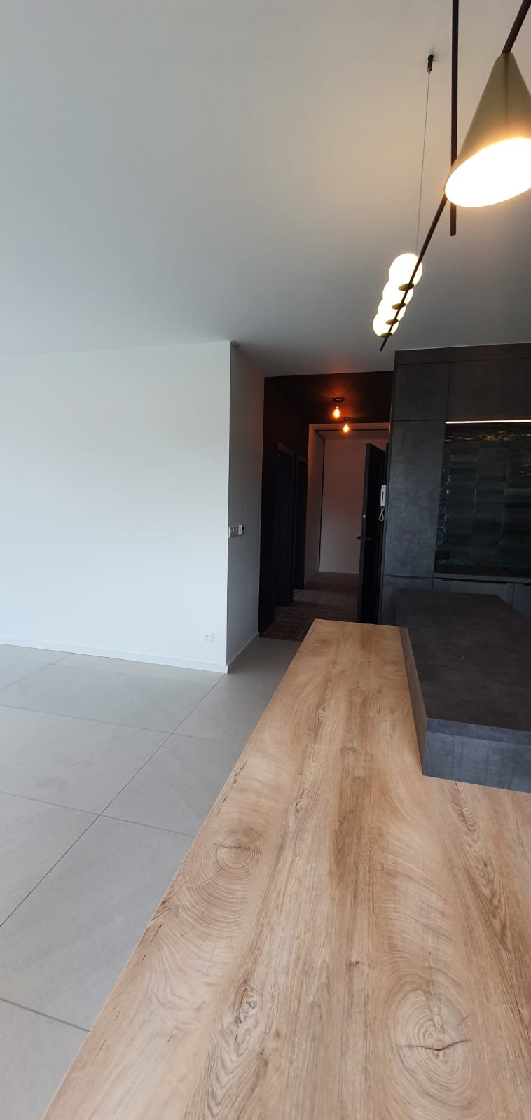 Nowoczesne wnętrze: jasne płytki podłogowe, białe ściany, drewniany blat wyspy kuchennej z widocznym rysunkiem słojów, ciemna zabudowa kuchenna i designerskie oświetlenie sufitowe.