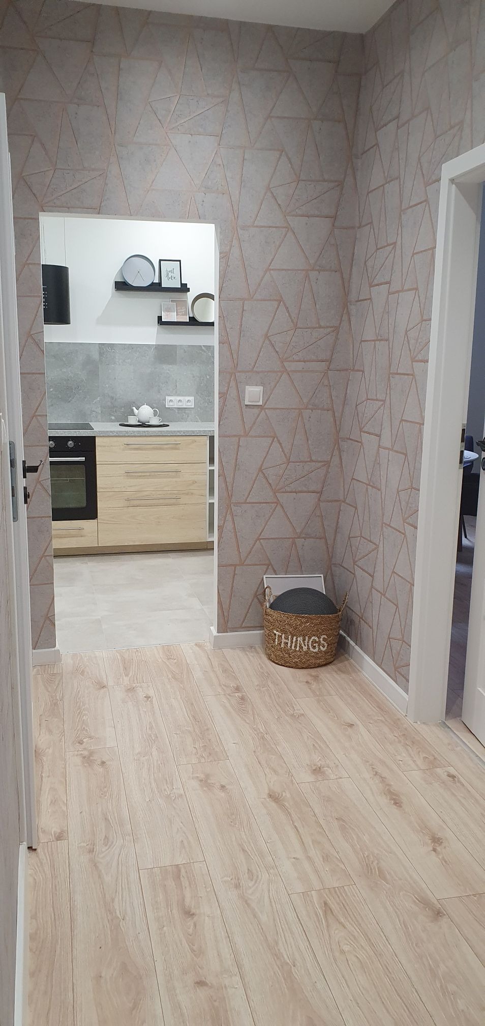 Wnętrze mieszkania z nową podłogą laminowaną, ścianą z geometryczną tapetą i widokiem na kuchnię z drewnianymi szafkami oraz czarną płytą grzewczą.