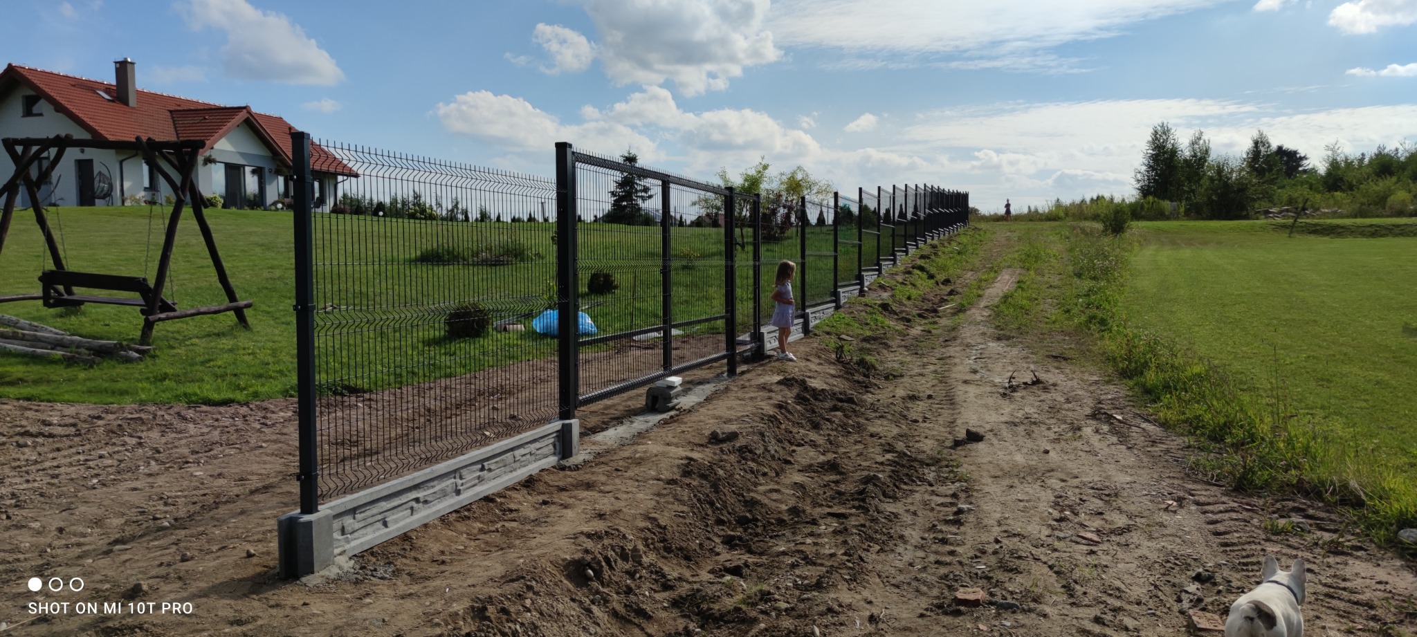 Długa linia panelowego ogrodzenia z betonową podmurówką, biegnąca wzdłuż polnej drogi, z małą dziewczynką przyglądającą się przez siatkę i psem w prawym dolnym rogu, w tle dom z czerwoną dachówką...