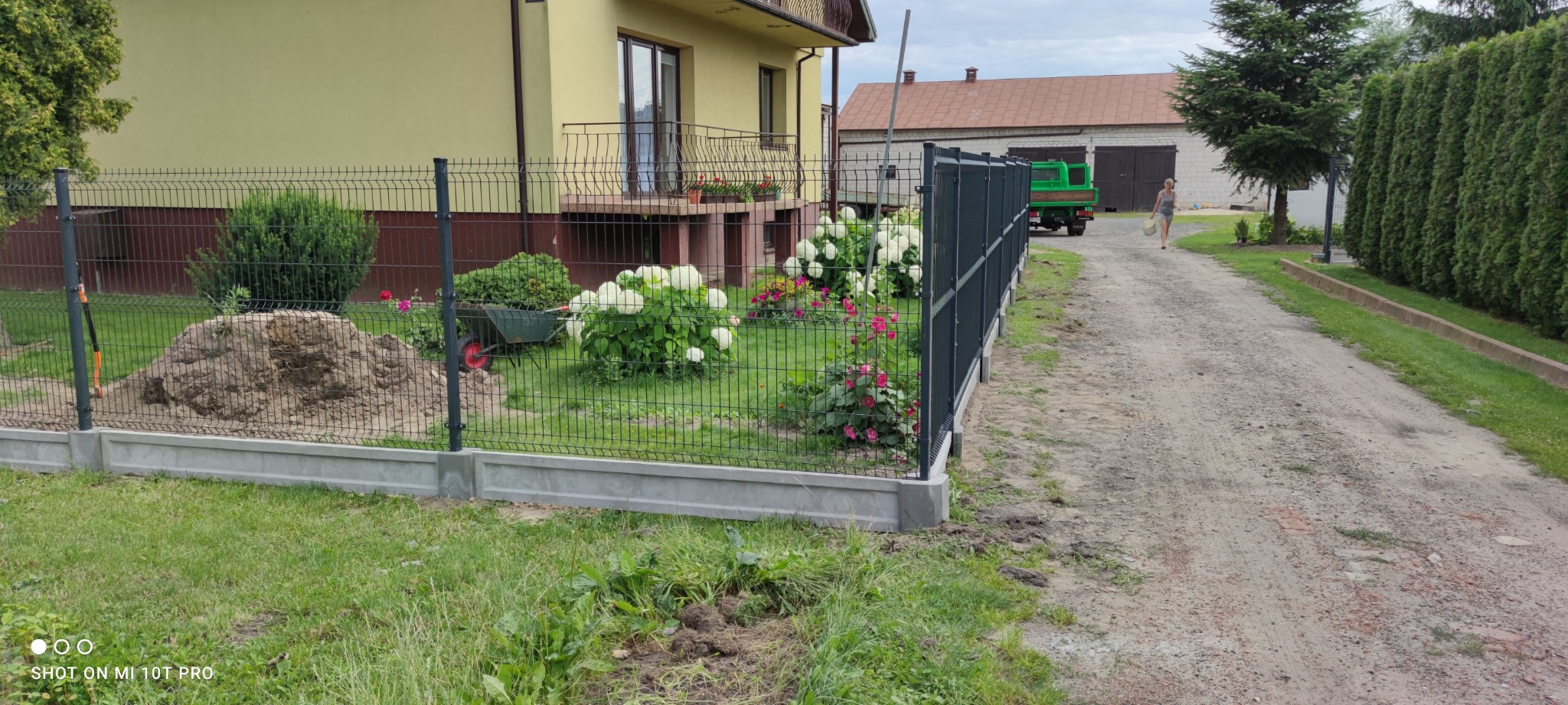 Nowo postawione, szare ogrodzenie panelowe na betonowej podmurówce, widok na ogród z hortensjami i różami, w tle dom z balkonem i zielony samochód dostawczy na żwirowej drodze.