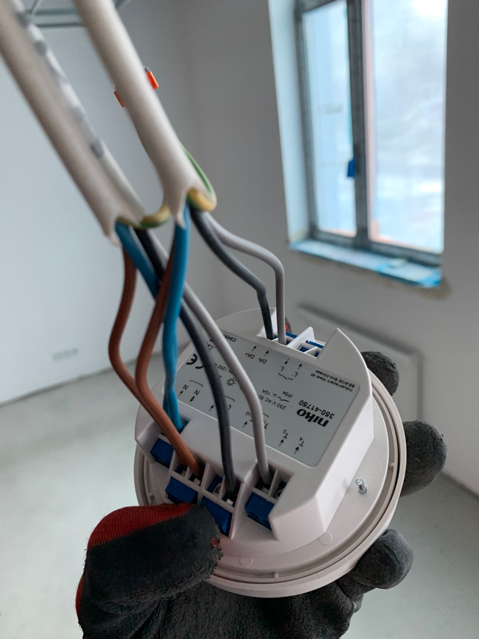 Elektryk w rękawicy podłącza przewody do okrągłego, białego modułu sterującego oświetleniem firmy Niko, widoczne oznaczenia i schematy na obudowie, w tle jasne wnętrze z oknem.