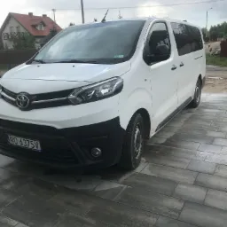 Biały bus Toyota Proace Verso zaparkowany na szarym bruku, widoczny numer rejestracyjny NO 6375V, w tle dom z czerwonym dachem i pochmurne niebo.