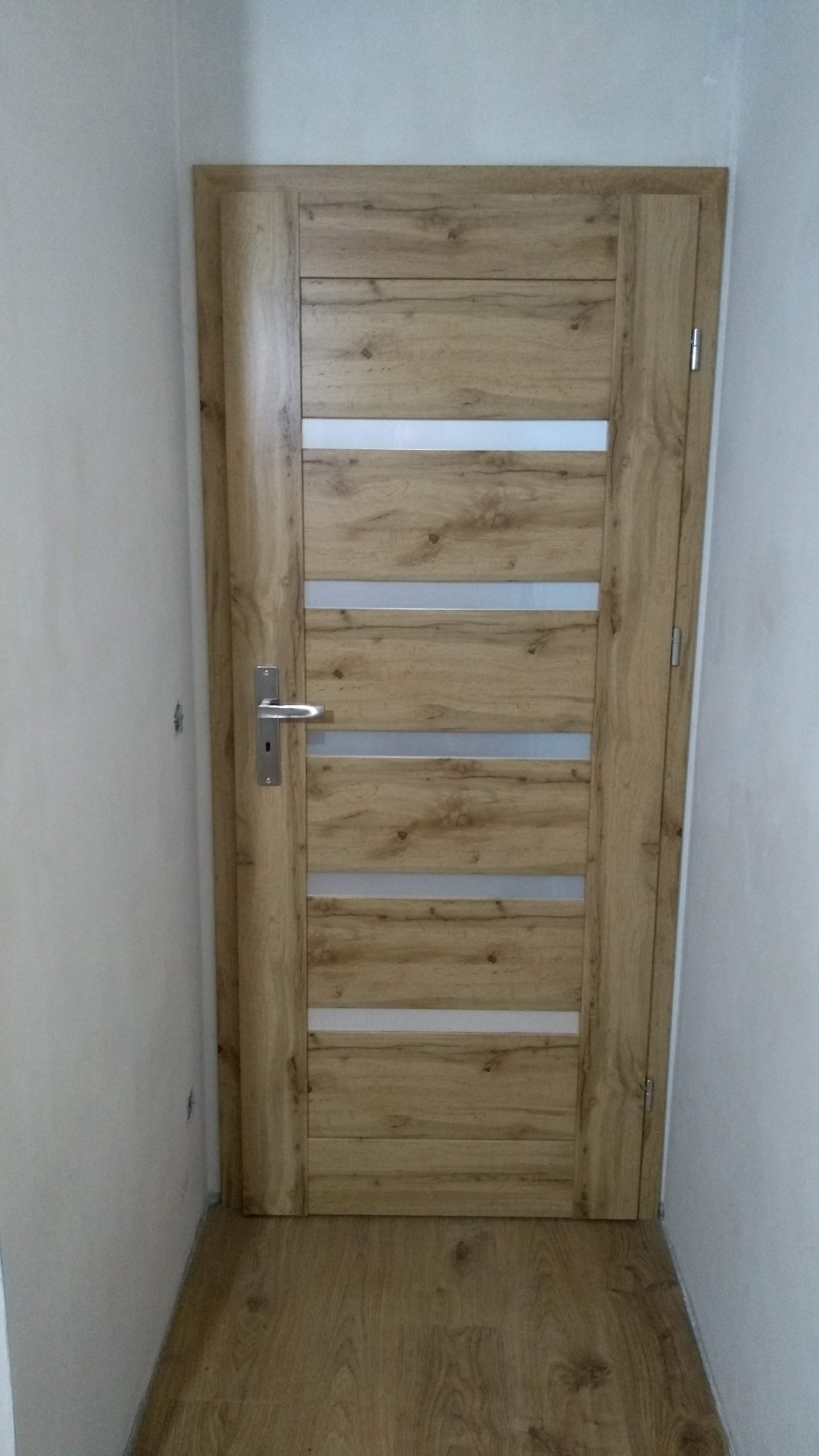 Zamknięte drzwi wewnętrzne z okleiną w kolorze jasnego drewna z czterema poziomymi, mlecznymi szybkami, srebrna klamka, widoczny fragment ściany i podłogi w odcieniach beżu.