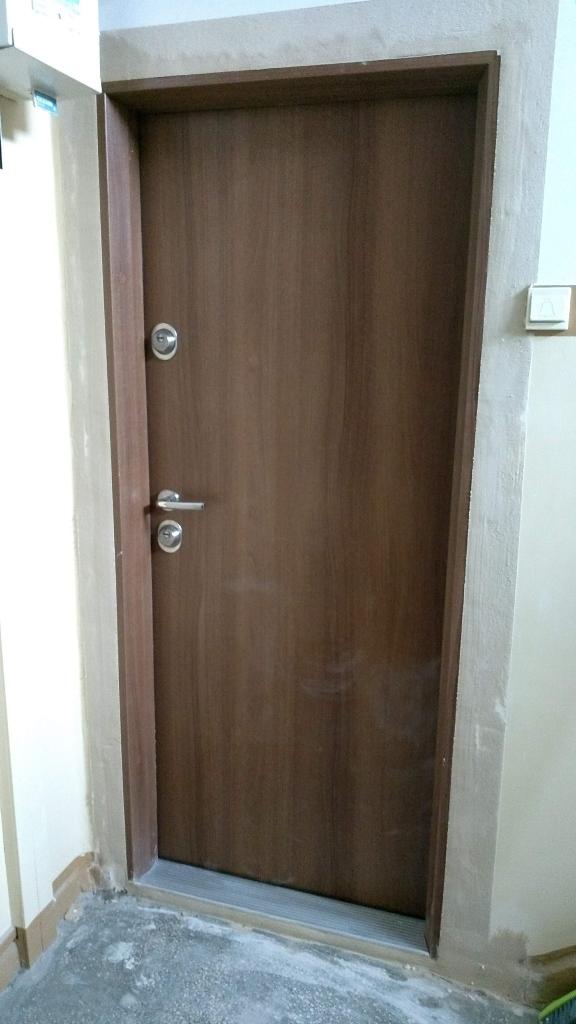 Brązowe drzwi wejściowe zamontowane w ościeżnicy, widoczny zamek z wkładką, klamka i dzwonek na ścianie.