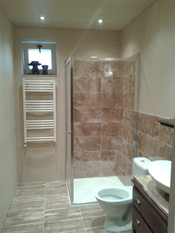 Wykończona łazienka z kabiną prysznicową z marmurkowymi płytkami, grzejnikiem drabinkowym pod małym oknem z figurkami, toaletą i umywalką nablatową na ciemnej szafce.