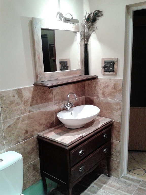 Umywalka nablatowa w stylu retro na ciemnej komodzie z dwiema szufladami, marmurowe płytki na ścianie, lustro w drewnianej ramie z półką, dekoracyjne pióra pawie w wazonie.