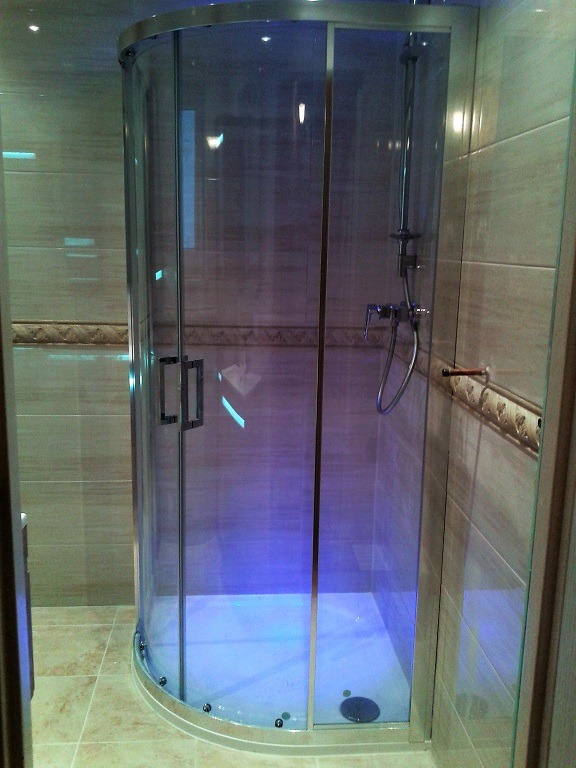 Zaokrąglona kabina prysznicowa z chromowanymi wykończeniami i transparentnymi ściankami, oświetlona niebieskim światłem, na tle beżowych płytek z dekoracyjnym paskiem.