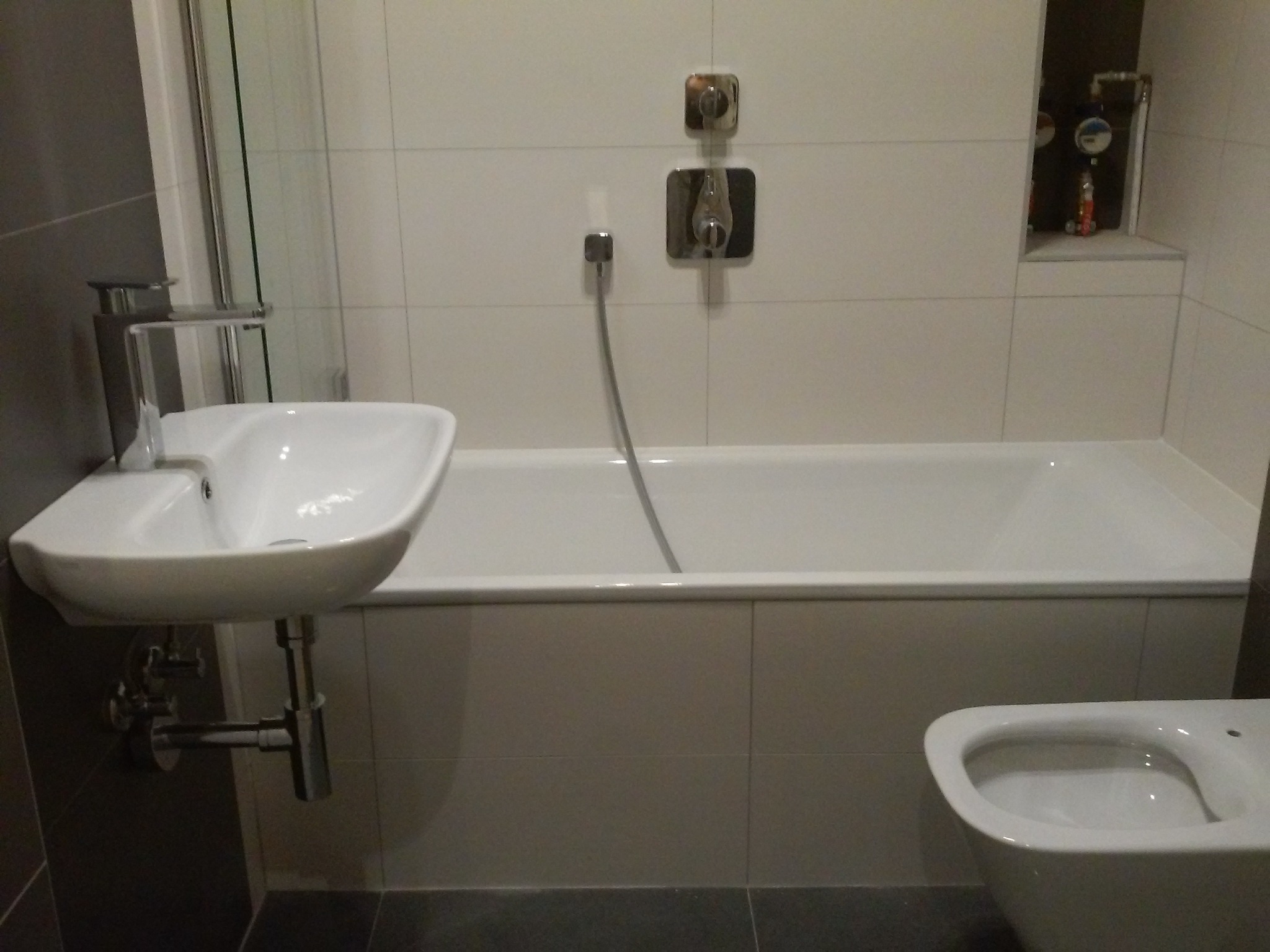 Wykończona łazienka z białą wanną, umywalką na ścianie z chromowaną baterią i wiszącą toaletą; na ścianie białe płytki kwadratowe, w niszy widoczne wodomierze.