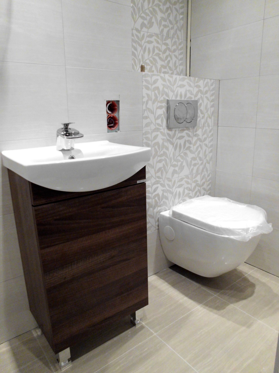 Nowoczesna łazienka z wiszącą toaletą w folii ochronnej, umywalką z szafką z ciemnego drewna na chromowanych nóżkach, oraz płytkami z motywem liści na ścianie, widoczna niezamaskowana puszka...