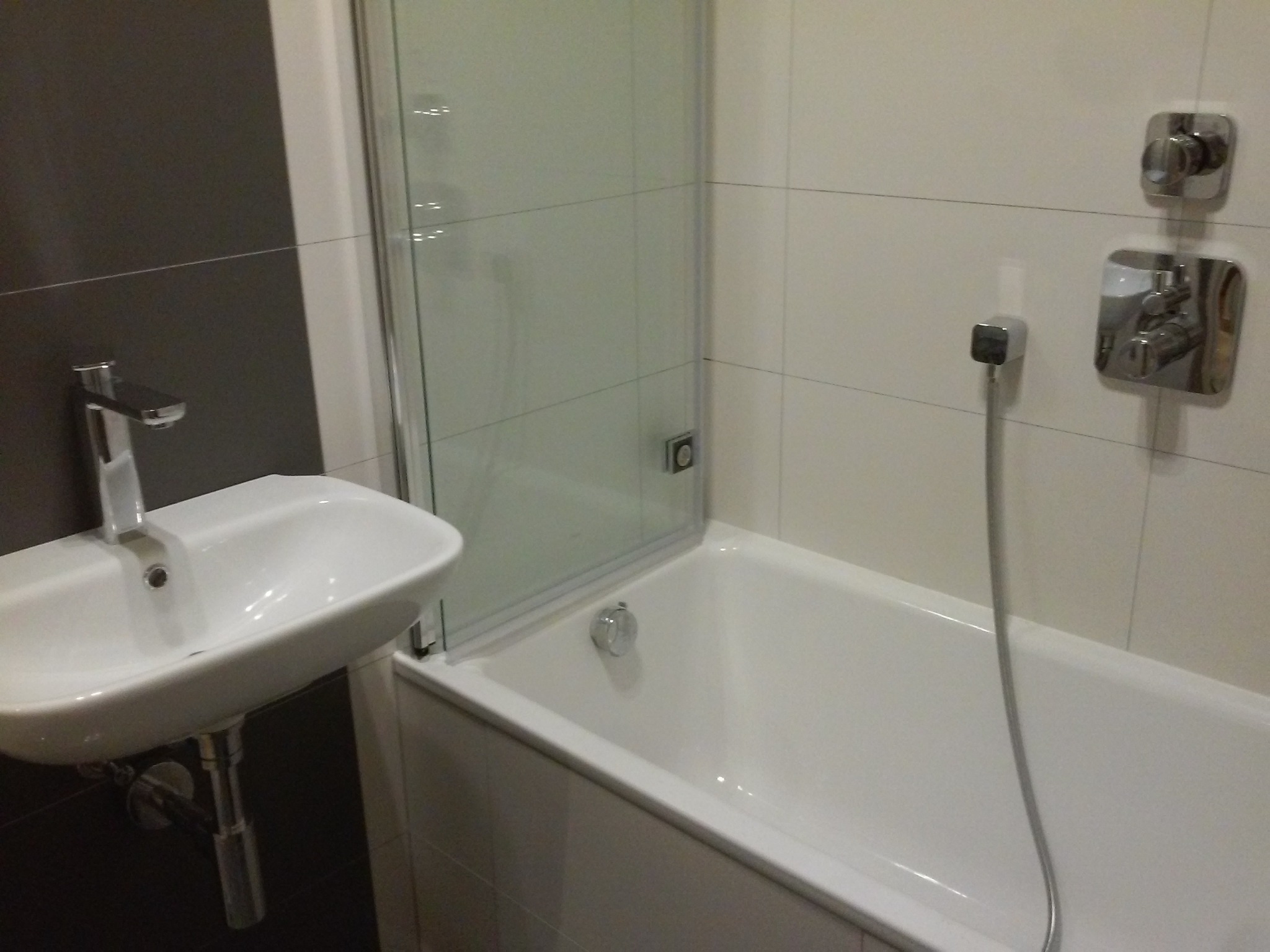 Wykończona łazienka z białą wanną z parawanem nawannowym, umywalką na postumencie i szarą ścianą z lustrzanym kranem.