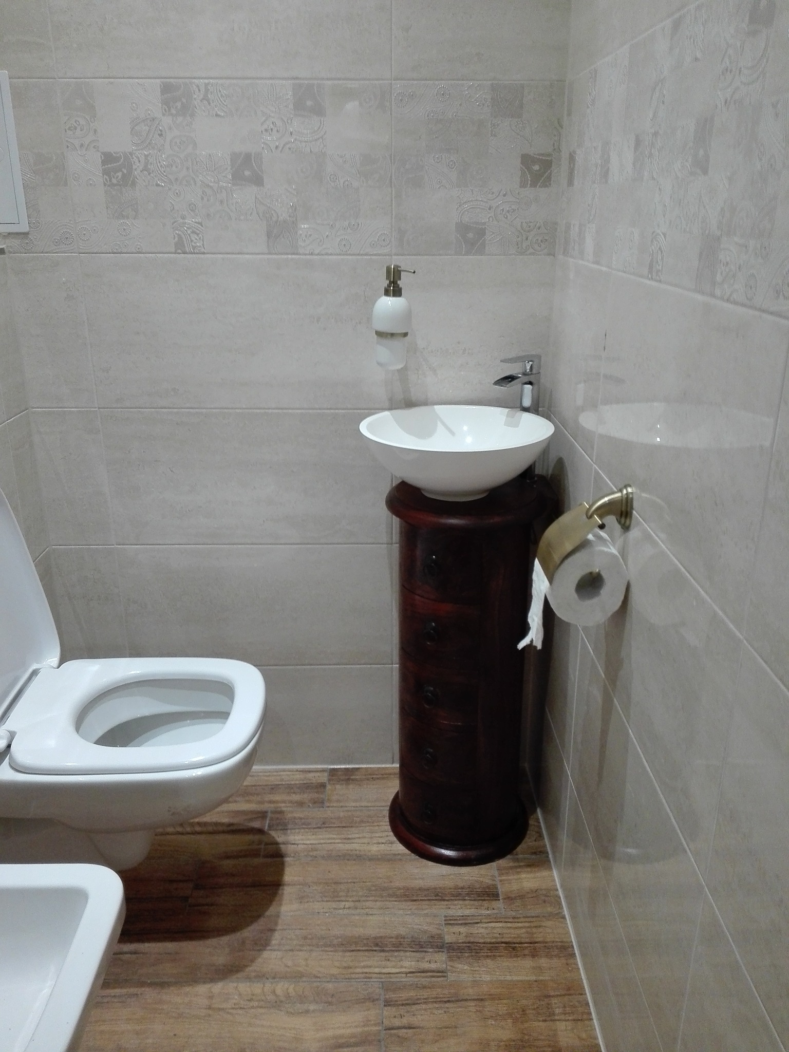 Wykończona łazienka z beżowymi płytkami ściennymi z delikatnym wzorem, białą toaletą i umywalką nablatową na ciemnobrązowej szafce z szufladami, drewnopodobna podłoga.