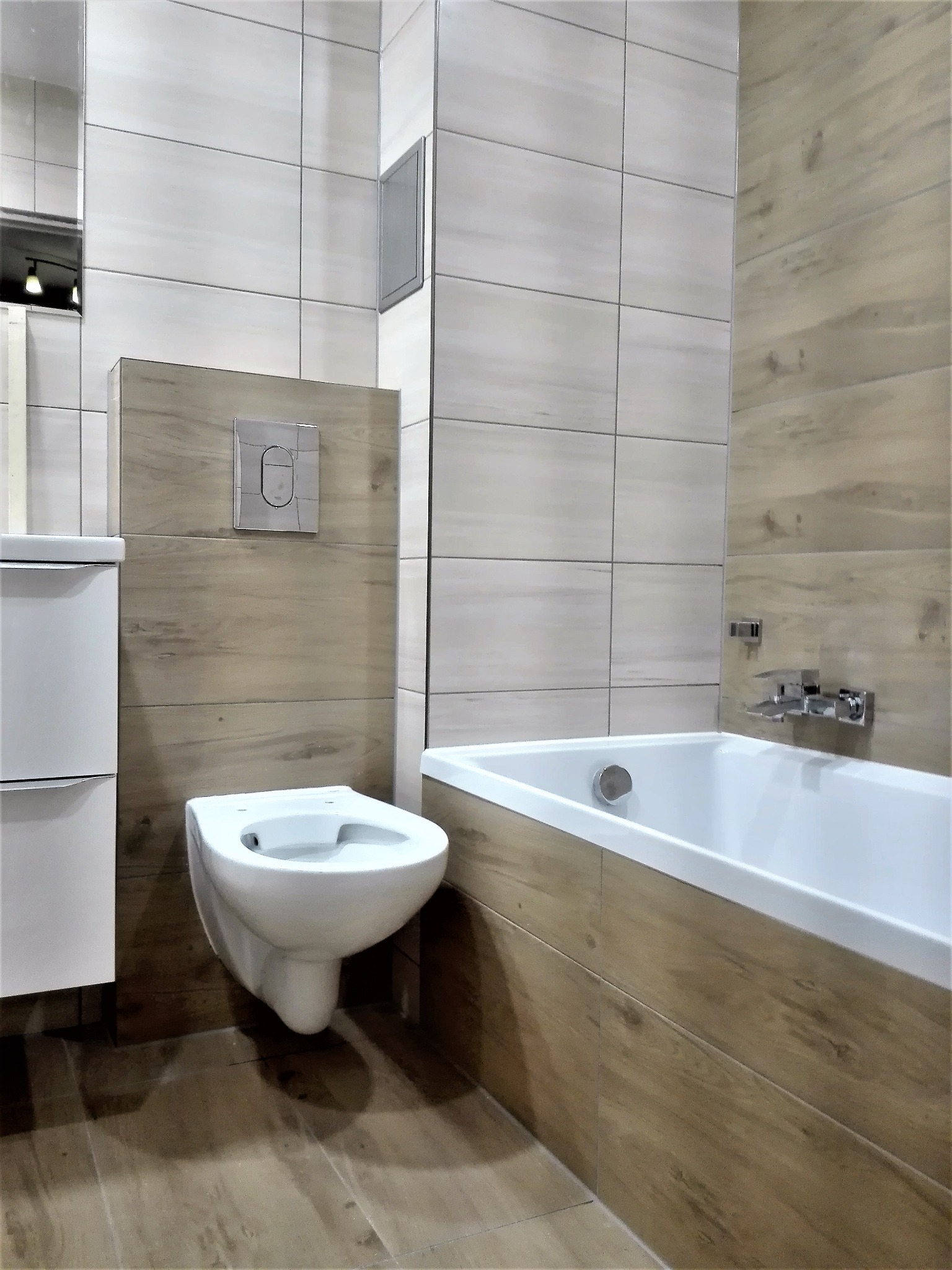 Nowoczesna łazienka z drewnopodobnymi płytkami, wisząca toaleta z chromowanym przyciskiem spłukiwania, biała wanna i umywalka z szafką.