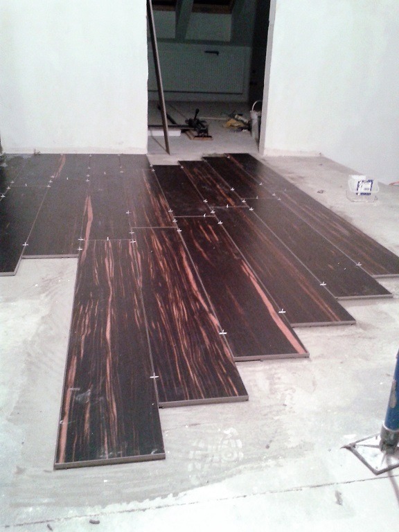 Układanie podłogi z płytek imitujących drewno, widoczne krzyżyki dystansowe, w tle narzędzia i wiadro z klejem.