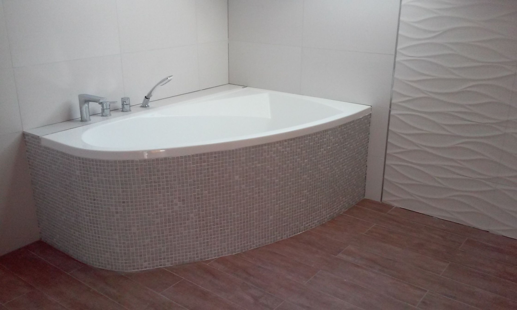 Narożna wanna obudowana mozaiką w łazience z panelami podłogowymi imitującymi drewno i dekoracyjną, falowaną płytą ścienną.