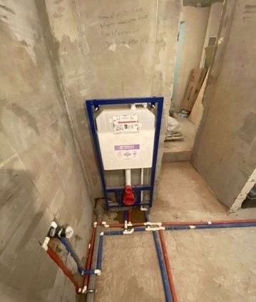 Stelaż podtynkowy WC zamontowany w surowym wnętrzu łazienki, widoczne rury doprowadzające wodę, ściany w stanie deweloperskim z widocznymi notatkami.