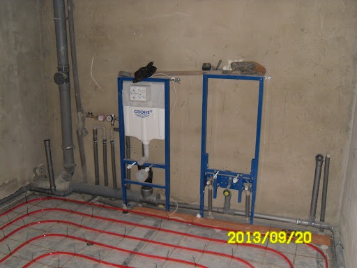 Surowa instalacja sanitarna w trakcie montażu: stelaż podtynkowy Grohe z widocznym zbiornikiem, rury kanalizacyjne i grzewcze podłogowe z czerwonego tworzywa, surowe ściany w tle.