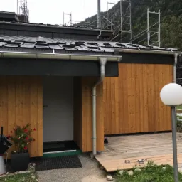 Nowoczesny budynek z drewnianą elewacją i ciemnym dachem z rynnami; w tle widoczne rusztowania i zalesione wzgórze.