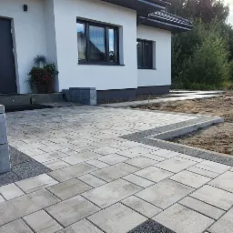 Nowoczesny podjazd z jasnoszarej kostki brukowej ułożonej wzorem prostokątnym, obrzeża z ciemnego grysu, rabata z ziemią przygotowana pod nasadzenia, biały dom z szarą stolarką okienną i drzwiową...