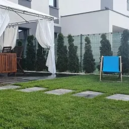 Zadbany ogród przydomowy z równo przystrzyżonym trawnikiem, ścieżką z kwadratowych płyt, dwoma niebieskimi leżakami, oraz zadaszoną altaną z białymi zasłonami, tworzący relaksującą przestrzeń.