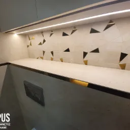 Wykończona łazienka z beżowymi płytkami o nieregularnym wzorze geometrycznym z elementami w kolorze złotym, podświetlana półka i zabudowa, widoczny przycisk spłuczki toalety.