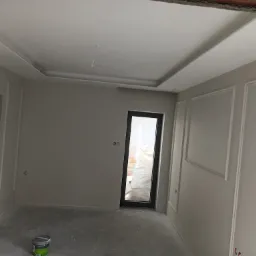 Wnętrze pokoju w trakcie prac wykończeniowych: ściany pomalowane na szaro z dekoracyjnymi listwami, sufit z ozdobnym obramowaniem, widoczna puszka z farbą na podłodze.
