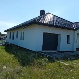 Jednorodzinny dom z szarą bramą garażową i świeżo pomalowaną białą elewacją, widok z zewnątrz w słoneczny dzień.