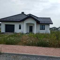 Jednorodzinny dom z białą elewacją, czarną dachówką i garażem, otoczony polną roślinnością i chodnikiem z kostki.