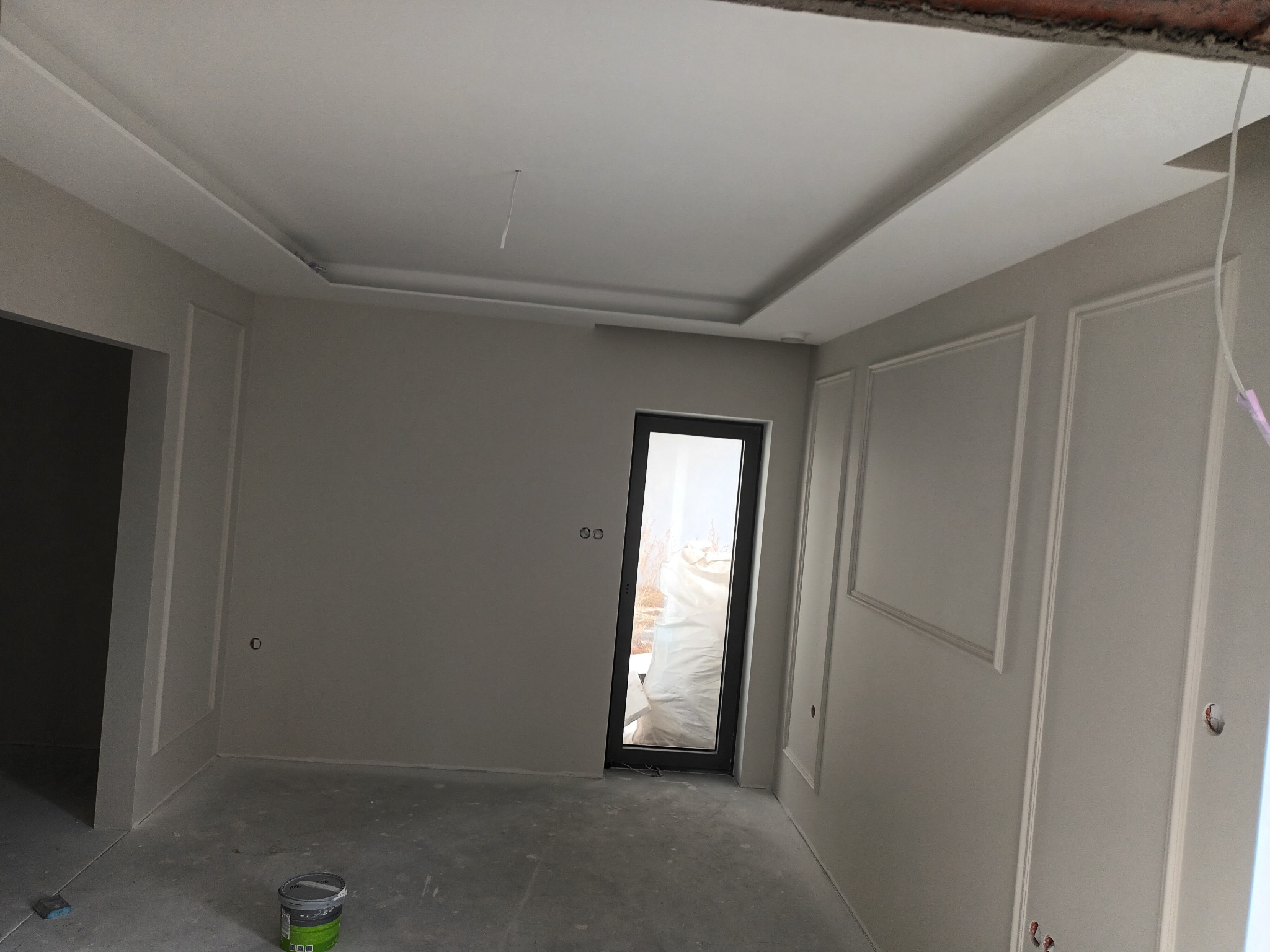 Wnętrze pokoju w trakcie prac wykończeniowych: ściany pomalowane na szaro z dekoracyjnymi listwami, sufit z ozdobnym obramowaniem, widoczna puszka z farbą na podłodze.