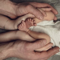 WAM Studio Fotografia - Dłonie rodziców otulają stopy noworodka na tle szarej, pikowanej kołdry.