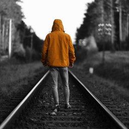 WAM Studio Fotografia - Osoba w pomarańczowej kurtce z kapturem i jasnych jeansach, stojąca tyłem na torach kolejowych, otoczona czarno-białym lasem.