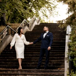 Elegancko ubrana para trzymająca się za ręce schodzi po betonowych schodach w parku, mężczyzna w granatowym garniturze z muchą, kobieta w białej sukience do kolan.