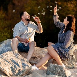 WAM Studio Fotografia - Para siedzi na kamieniach na plaży, mężczyzna puszcza bańki mydlane, kobieta wyciąga rękę, by je dotknąć, ciepłe światło zachodzącego słońca