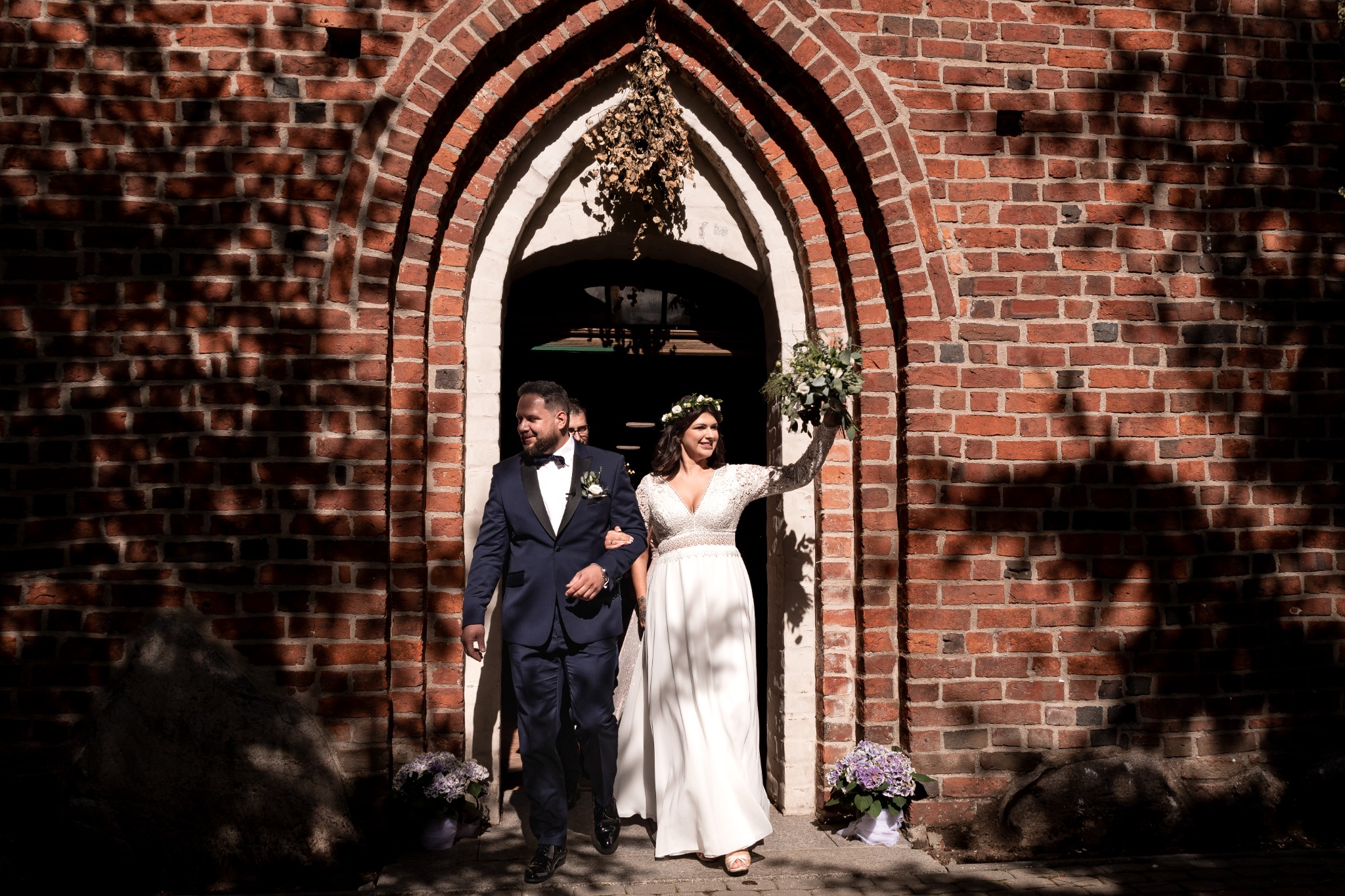 Para młoda wychodzi z kościoła z czerwonej cegły, panna młoda trzyma bukiet kwiatów, cień drzewa na ścianie.