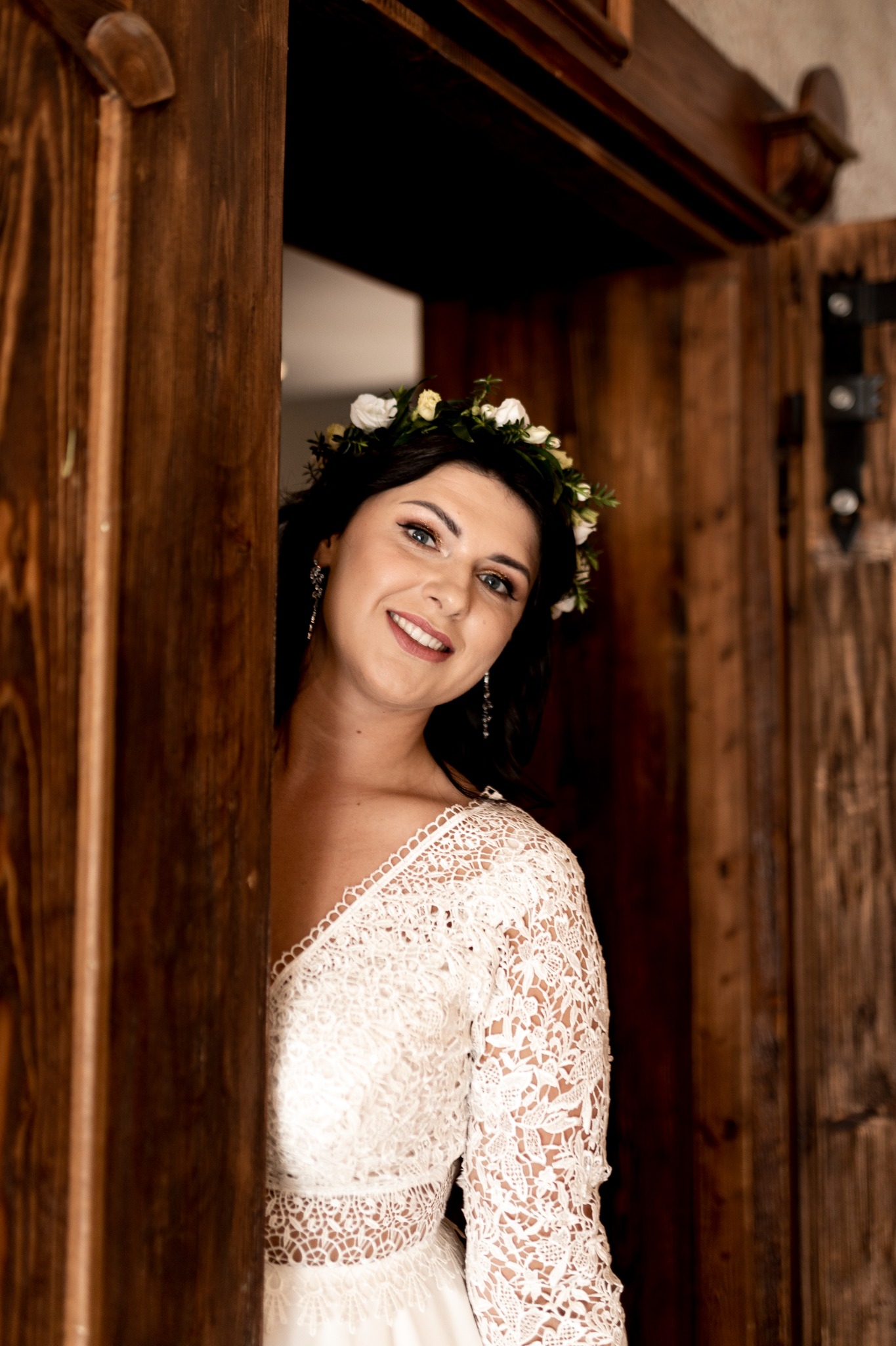 Uśmiechnięta panna młoda w koronkowej sukni i wianku z białych kwiatów, spoglądająca zza drewnianych drzwi, z delikatnym makijażem podkreślającym niebieskie oczy.