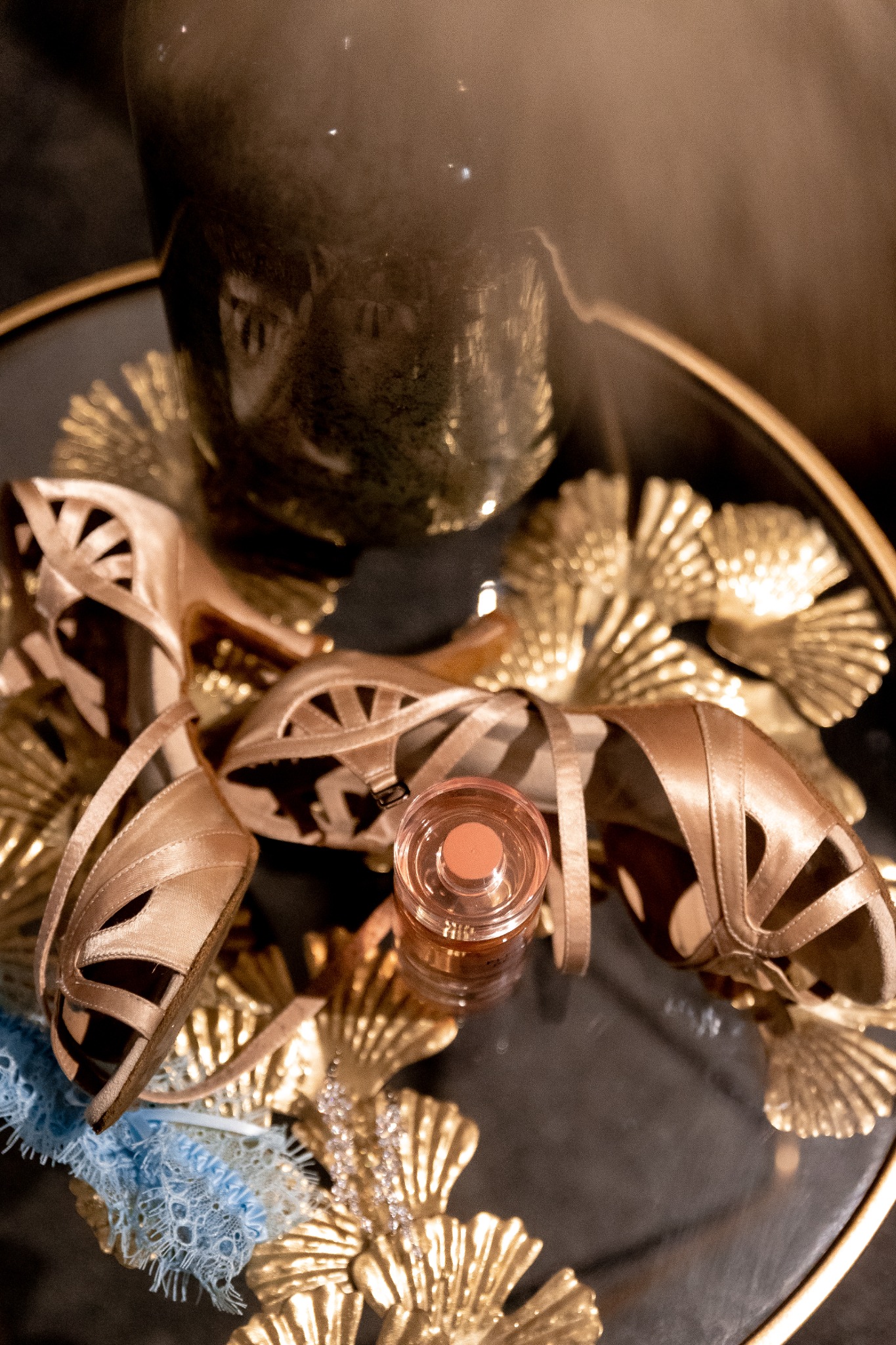 Beżowe, eleganckie buty damskie na wysokim obcasie, mała buteleczka perfum i błękitna podwiązka na czarnym, okrągłym stoliku z dekoracjami w kolorze złota.