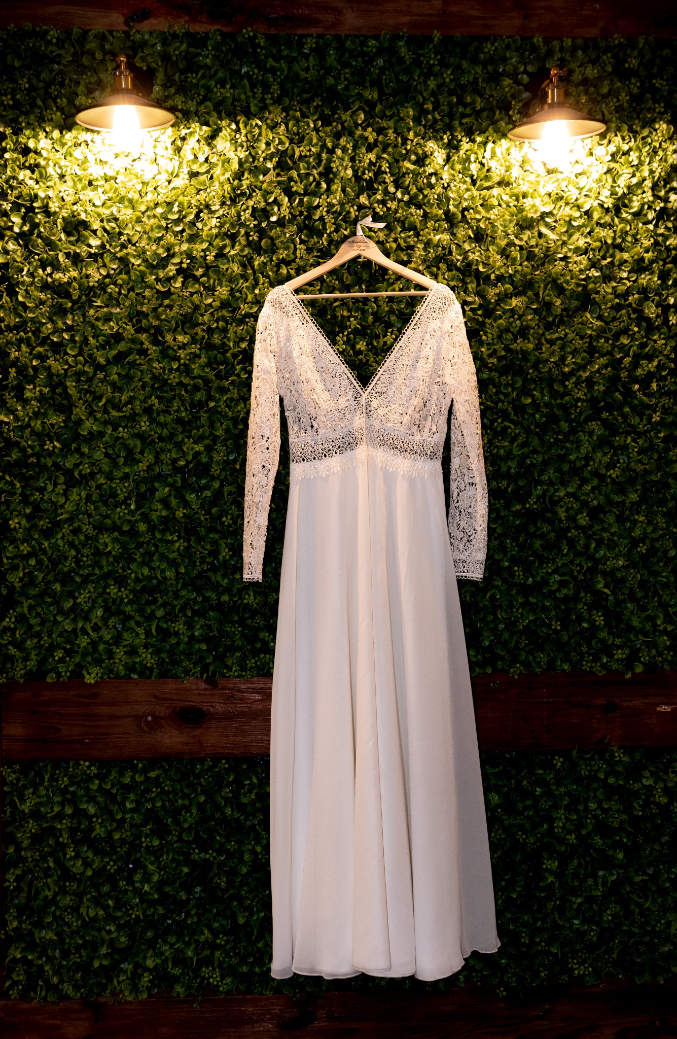 Elegancka suknia ślubna z koronkową górą i długimi rękawami, zawieszona na drewnianym wieszaku na tle ściany z zieleni, oświetlona ciepłym światłem lamp.