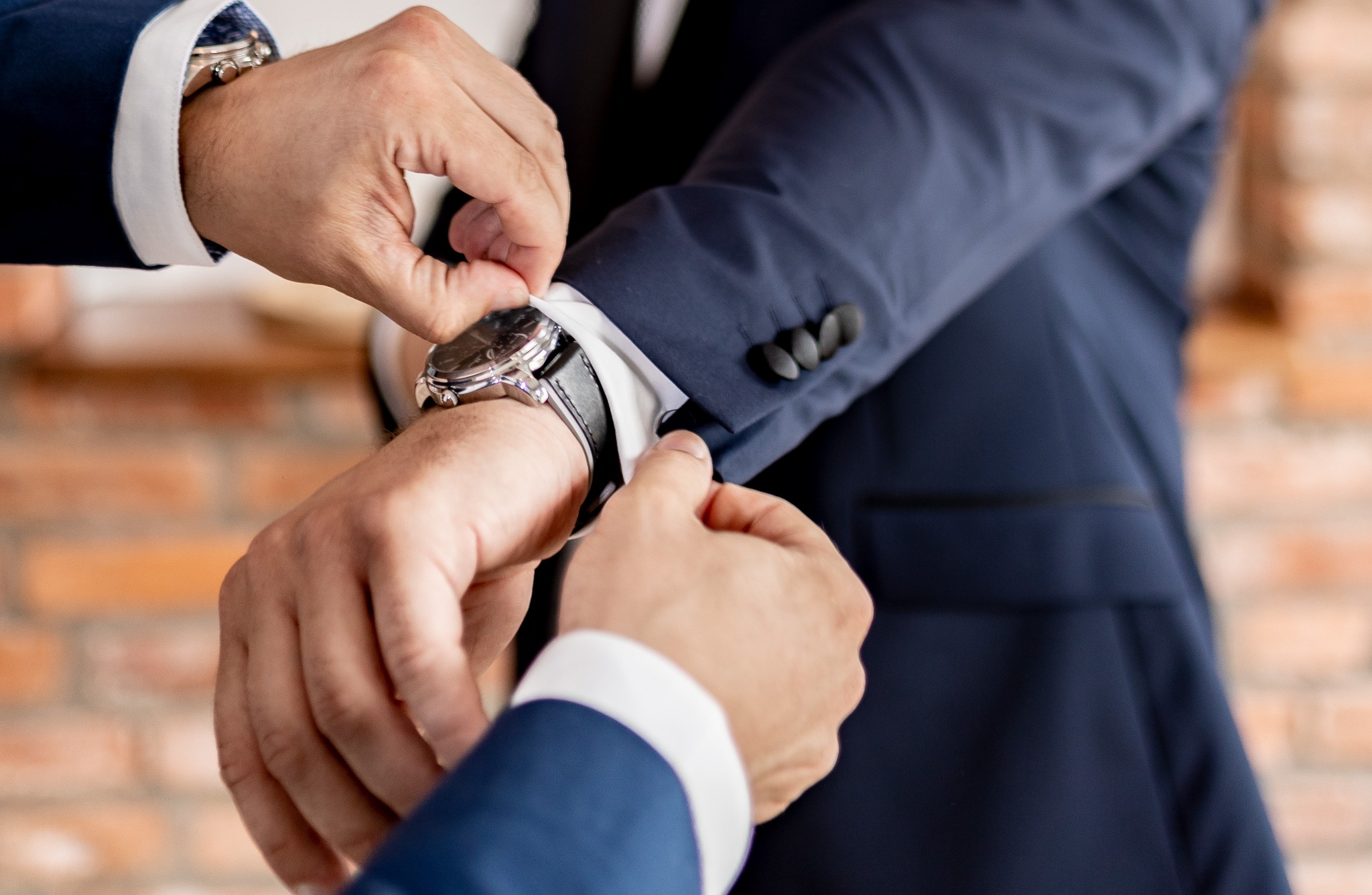 Mężczyzna zapinający mankiet granatowej marynarki, widoczny zegarek na skórzanym pasku, ceglana ściana w tle.
