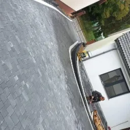 Świeżo ułożona, ciemnoszara kostka brukowa z białymi obrzeżami, widoczna zagęszczarka wibracyjna i pracownik w trakcie wykończenia, na tle domu z czerwoną dachówką.