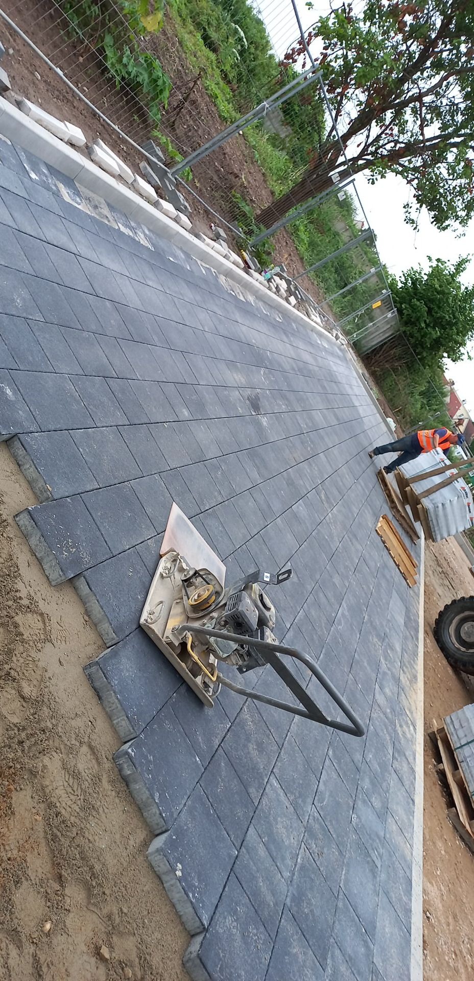 Proces zagęszczania ułożonej, szarej kostki brukowej za pomocą zagęszczarki wibracyjnej, widoczny pracownik w kamizelce odblaskowej i palety z kostką w tle.