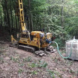 Żółta wiertnica na gąsienicach, ustawiona na betonowych płytach w zalesionym terenie, z podłączonymi zbiornikami na wodę, przygotowana do wiercenia.